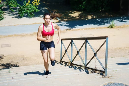 An athletic woman in activewear is focused and determined, running up stairs in a sunny park. Its part of her fitness routine, showing her dedication to active and healthy lifestyle