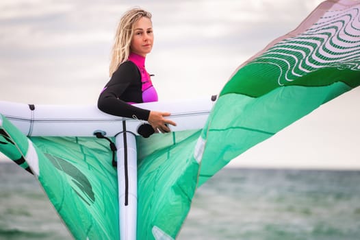 Portrait of a young female kitesurfer in a wetsuit holding her kite and looking at the camera against the background of the sea and sky.