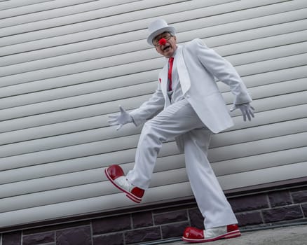 An elderly man in a white suit, huge boots and a clown nose walks funny