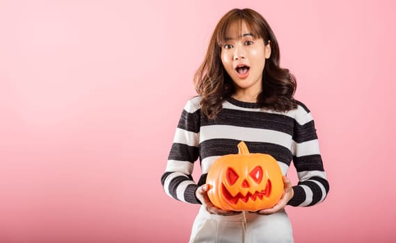 Portrait of a happy Asian woman in a Halloween costume, holding model pumpkins, including a ghost-shaped one. The studio shot on pink background captures the Halloween fun and excitement.