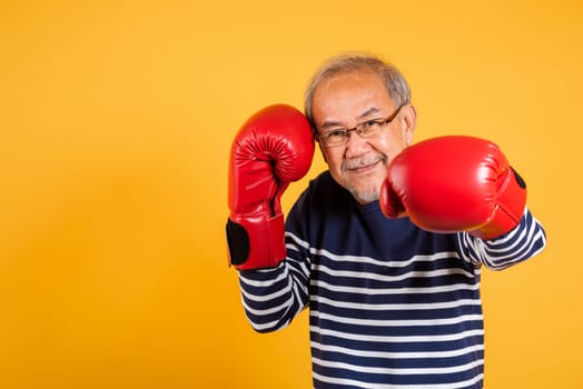 Portrait Asian old man wearing glasses wear two red boxing gloves studio shot isolated yellow background, smiling happy elderly man gray haired healthy fighter lifestyle concept