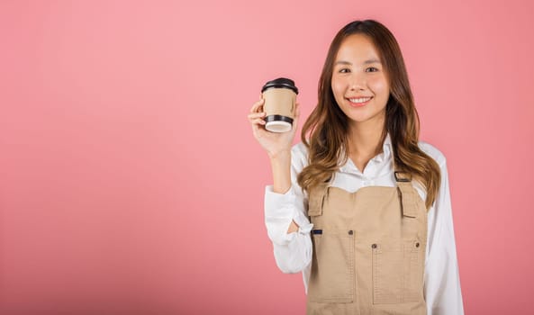 Portrait of Asian happy young woman barista bar tender coffee maker holding coffee tea hot cup studio shot isolated on pink background, female person smiling hold takeaways cup, small business