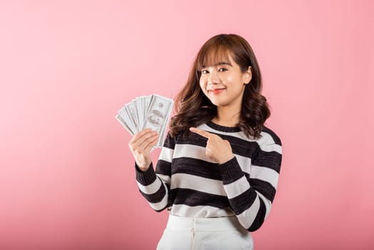 Smiling face portrait asian beautiful woman cheerful her holding and point the money fan banknotes 100 dollar bills on pink background, with copy space for text