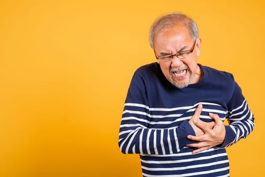 Portrait Asian old man with glasses holds hands on chest studio shot isolated yellow background, senior man pensioner with grey hair heart disease suffering from chest pain