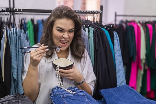 Happy fat woman fooling around and pretending to eat a pearl necklace with Chinese chopsticks in a clothing store for large people