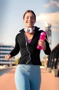 A young fitness girl runs with headphones and water bottle outdoors. Concept of workouts and healthy lifestyle.