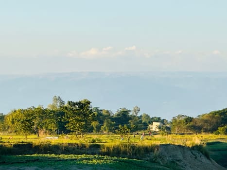 A scenic rural landscape with a distant mountain range in the background. The foreground features a field with people working, possibly tending to crops. The sky is clear and bright, with a few wispy clouds. The image captures the tranquility and beauty of the countryside.