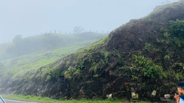 A scenic view of a misty mountainside with lush vegetation and a person standing near a road, taking in the beautiful scenery. The fog blankets the top of the hill, creating a sense of mystery and tranquility.