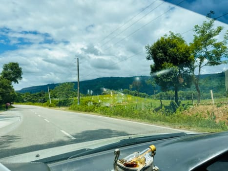 A view from inside a car as it drives along a paved road surrounded by lush green landscapes. The road is straight and the sky is a bright blue with fluffy white clouds. There are green trees and hills in the distance. The photo was taken on a sunny day.