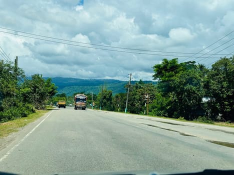 A rural road cutting through a lush green landscape with rolling hills in the background. The road is lined with trees and vegetation, and the sky is dotted with white clouds. Two vehicles can be seen driving on the road, one in the distance and one in the foreground. The image evokes a sense of tranquility and peacefulness.