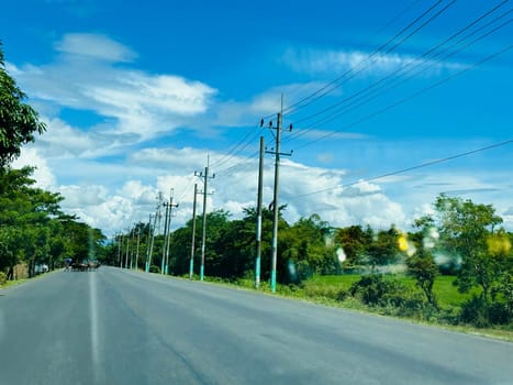 A wide, paved road stretches out straight ahead under a bright blue sky with fluffy white clouds. The road is bordered by green trees and lush foliage, with power lines running along the side. The sun is shining brightly, casting a warm glow on the scene.