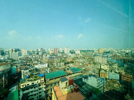A panoramic view of the cityscape of Dhaka, Bangladesh, on a sunny day. The image shows a dense urban environment with buildings of varying heights. The sky is clear and blue, with a few white clouds. The buildings are mostly brown and gray, with some green vegetation visible.