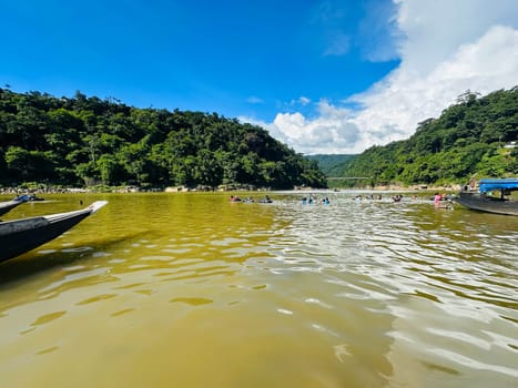 A view of a river in Bangladesh from the front of a boat. The water is calm and brown, reflecting the bright blue sky and fluffy white clouds. Green hills line the riverbanks and a bridge can be seen in the distance. People swim and travel by boat in the river.