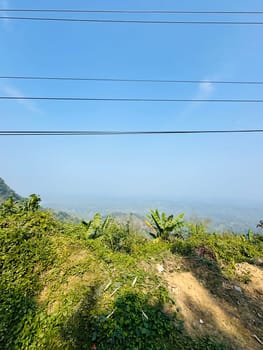 A scenic view from a hillside, showcasing lush greenery and a hazy landscape stretching out in the distance. Several power lines run across the clear blue sky above. The scene is bathed in the soft light of a sunny day.