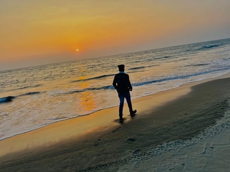 A man walks along a sandy beach towards the ocean as the sun sets, casting a golden reflection in the water. The sky is a warm orange and the water is a light blue.