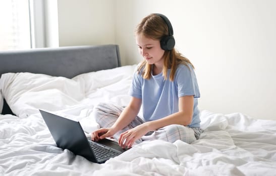 Sweet Young Girl Sitting In Bed, Putting On Headphones And Opening Her Laptop To Do Her Homework Online