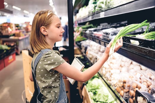 Pretty girl child buying green onion with shopping list in supermarket. Beautiful female preteen kid choosing vegetables in grocery store