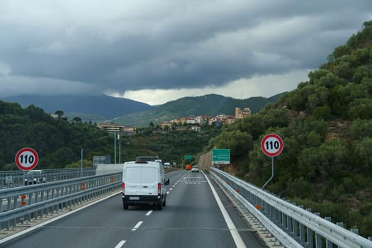 Castellaro, Italy - May 12, 2023: A white van travels on a highway bridge with a speed limit sign indicating 110 kilometers per hour. The bridge overlooks a town nestled between green mountains and the sky is cloudy.