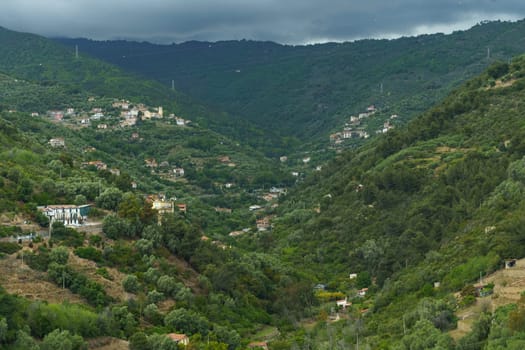 A scenic view of green hillsides dotted with houses under a cloudy sky. The image captures the beauty of a Mediterranean landscape with its rolling hills, dense foliage, and scattered settlements.