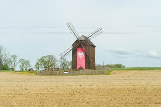 A solitary traditional windmill with red details stands in a plowed field, under a cloudy sky.