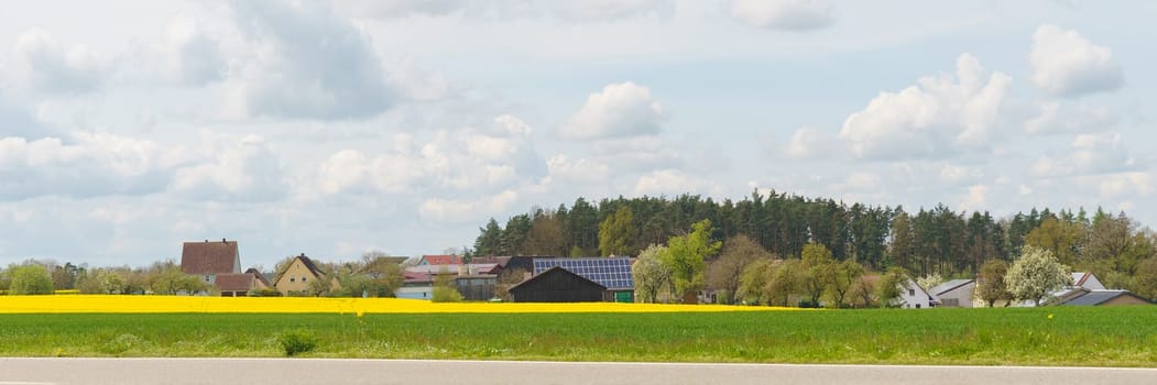 A scenic view of a rural village nestled amongst rolling fields and a forest, with a vibrant yellow field in the foreground. The sky is filled with fluffy white clouds, creating a tranquil atmosphere.