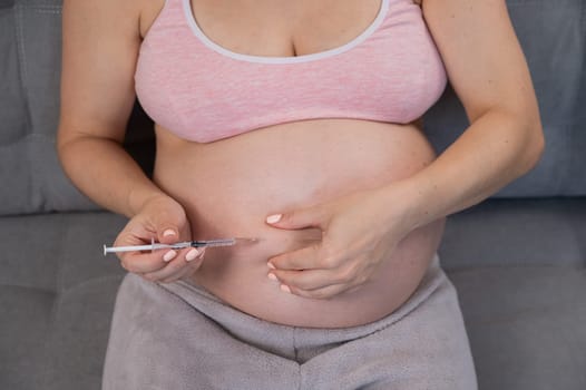 A pregnant woman puts an injection of insulin while sitting on the couch. Close up of the belly