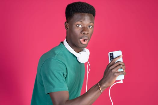 Young black man looking surprised with black headphones plugged into a mobile phone on a red background, with copy space.