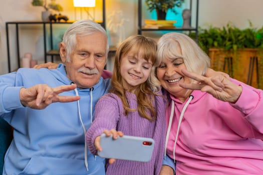Happy grandfather, grandmother and granddaughter taking selfie photo with smartphone at home. Smiling Caucasian girl with grandparents make blogger self portrait showing peace sign sitting on sofa.