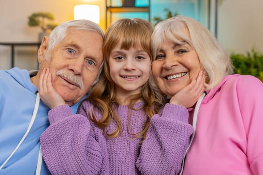 Portrait of smiling family kid granddaughter and loving grandparents looking at camera. Loving girl child with grandfather and grandmother embracing sitting on sofa in room at home. Love bonding care