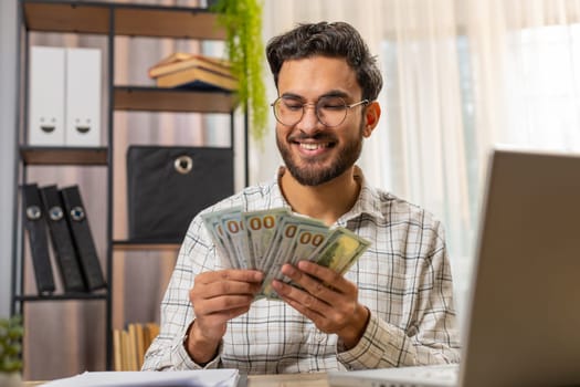 Happy young Indian businessman counting money cash and using laptop in home office. Successful Arabian freelancer getting bundle of money in office, calculates dividends cash money profits finances.