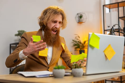 Tired exhausted frustrated bearded businessman sits in home office table with laptop sticky notes with many tasks inscription help, feels sad boredom overworked multitasking. Red hair guy freelancer.