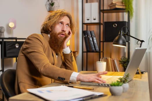 Bored sleepy bearded businessman worker working on laptop computer, leaning on hand at home office desk. Exhausted tired Caucasian red hair freelancer workaholic guy. Employment, occupation, workless