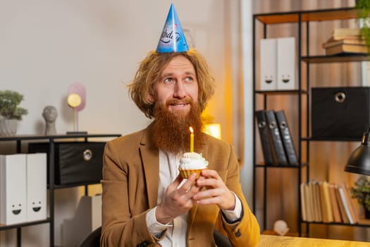 Happy Caucasian businessman wears festive birthday hat hold cupcake makes wish joyful congratulating blowing burning candle on cupcake. Redhead man freelancer celebrating birthday party at home office