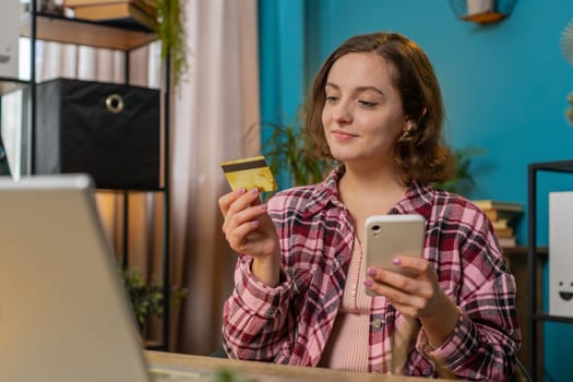 Happy Caucasian woman using bank credit card and smartphone for online shopping payments at home table. Business transaction. Smiling girl freelancer purchases with cellphone E-banking app service.