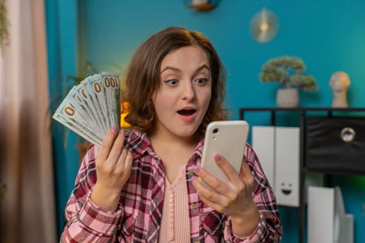 Rich excited amazed shocked young woman freelancer looks at camera holding money cash and smartphone. Female businesswoman girl satisfied of income earnings saves money sitting at home office at table