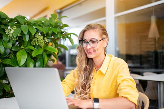 Woman freelancer wearing glasses and yellow shirt sitting at table, using laptop computer. Houseplant on background. Smiling businesswoman working with laptop sitting cafe terrace