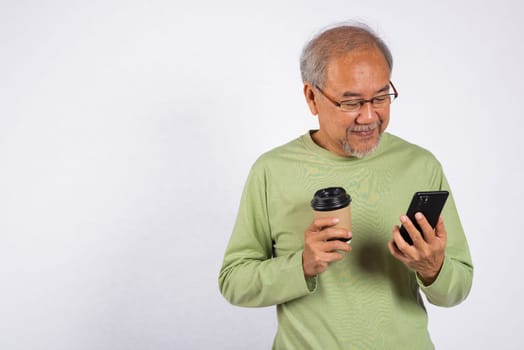 Portrait Asian old man while holding a cell phone and a cup of coffee studio shot isolated on white background. Concept of relaxation and enjoyment, elderly man is enjoy his coffee and check his phone