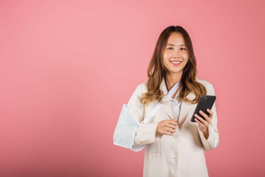 Woman confident smiling broken arm after accident and wear arm splint for treatment and hold smartphone, happy Asian female sling supported hand isolated on pink background, social media, copy space