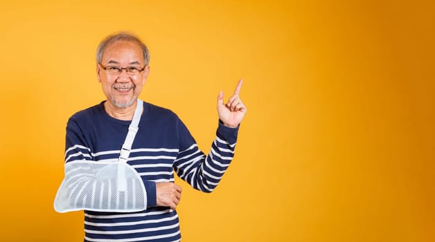 Portrait Asian old man cast on his arm is pointing space studio shot isolated yellow background, retired elderly man after accident broken arm wear arm splint for treatment accident insurance concept