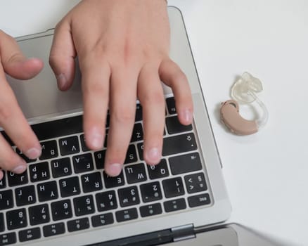 Close-up of a man's hands on a laptop keyboard. Hearing aid on the desktop