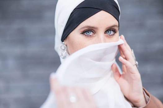 Portrait of a young blue-eyed woman in a hijab against a gray brick wall. Muslim woman with white scarf covering her face