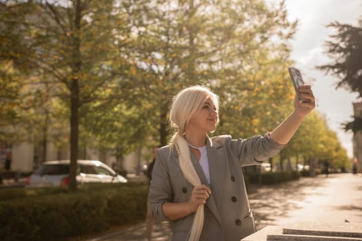 A woman is taking a picture of herself with her cell phone. She is wearing a gray jacket and scarf. The scene is set in a city with trees and cars in the background