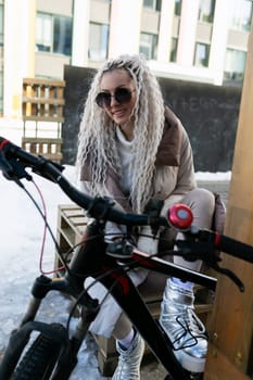 A woman is seated on a bench next to a bicycle. She appears to be resting or taking a break from cycling. The bike is parked next to her on the sidewalk.