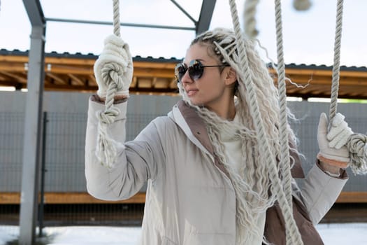A woman wearing sunglasses and a scarf is seated on a swing, gently swaying back and forth. The swing is suspended from a sturdy frame, and the womans expression is relaxed.