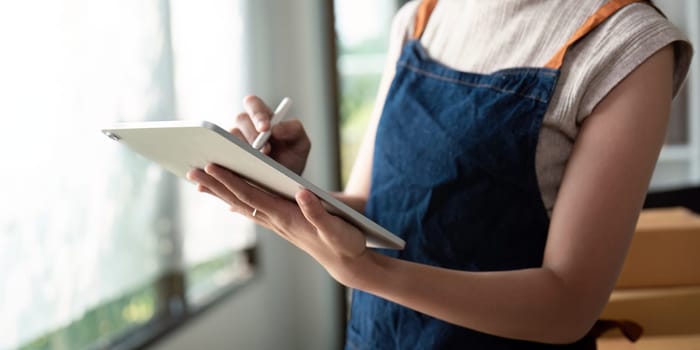 A young entrepreneur in an apron uses a digital tablet to manage and prepare parcel boxes for online ecommerce business and product delivery.