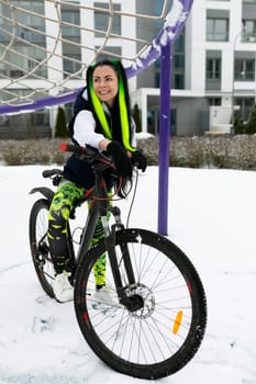 A woman is seated on top of a stationary bike in a snowy landscape. She appears to be taking a break from riding and is bundled up in winter clothing.