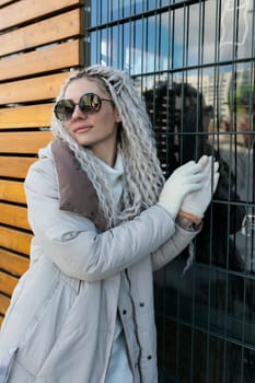 A woman wearing a white coat and sunglasses leans casually against a solid wall. She exudes a sense of confidence and poise as she stands in a relaxed pose.