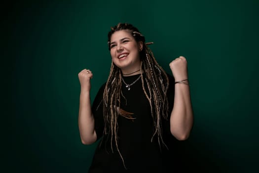 A woman with dreadlocks is standing against a solid green background. She appears confident and relaxed, looking directly at the camera.