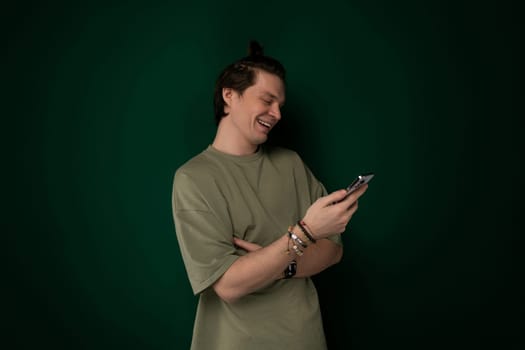 A man is standing in front of a vibrant green wall, holding a cell phone in his hand. He appears to be engaged with the device, possibly texting or browsing. The man is wearing casual attire and seems focused on his phone.