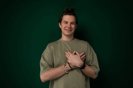 A man is standing upright in front of a solid green wall. He appears to be looking straight ahead, with a neutral expression on his face. The wall provides a simple background for the mans presence.